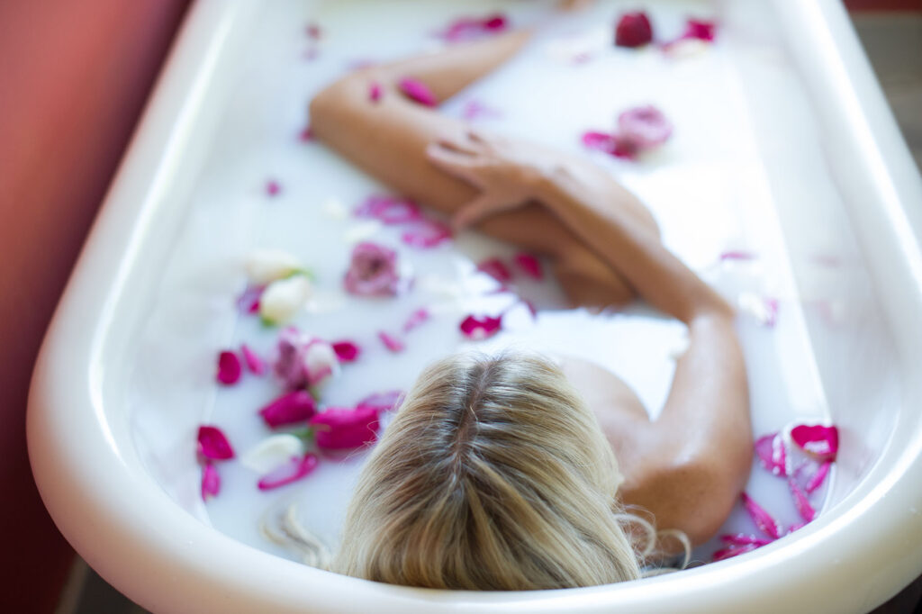 A person relaxes in a milky bath surrounded by pink and red rose petals. The serene setting, with their arms crossed, conveys tranquility and relaxation.