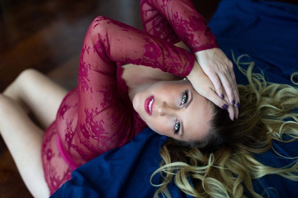 Woman in red lace dress lies on blue fabric, gazing upward. Her hands rest on her head, and long blonde hair fans out, creating a serene, intimate atmosphere.