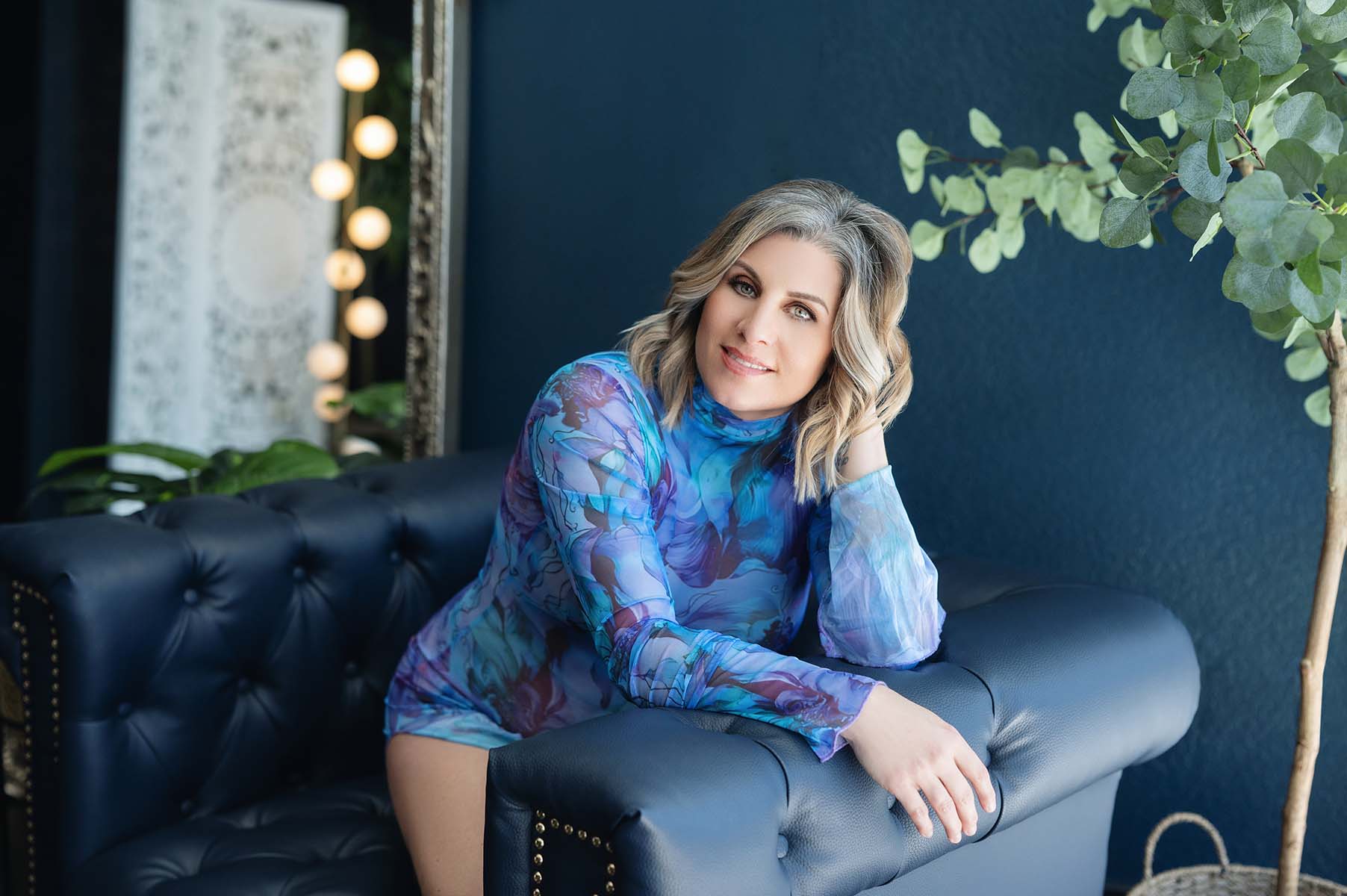 Cool toned boudoir portrait of blonde woman wearing blue floral long sleeve bodysuit kneeling on blue tufted couch