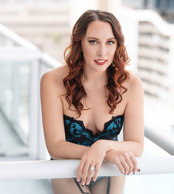 Brightly lit portrait of woman posing outdoors on a balcony wearing a blue and black lace corset