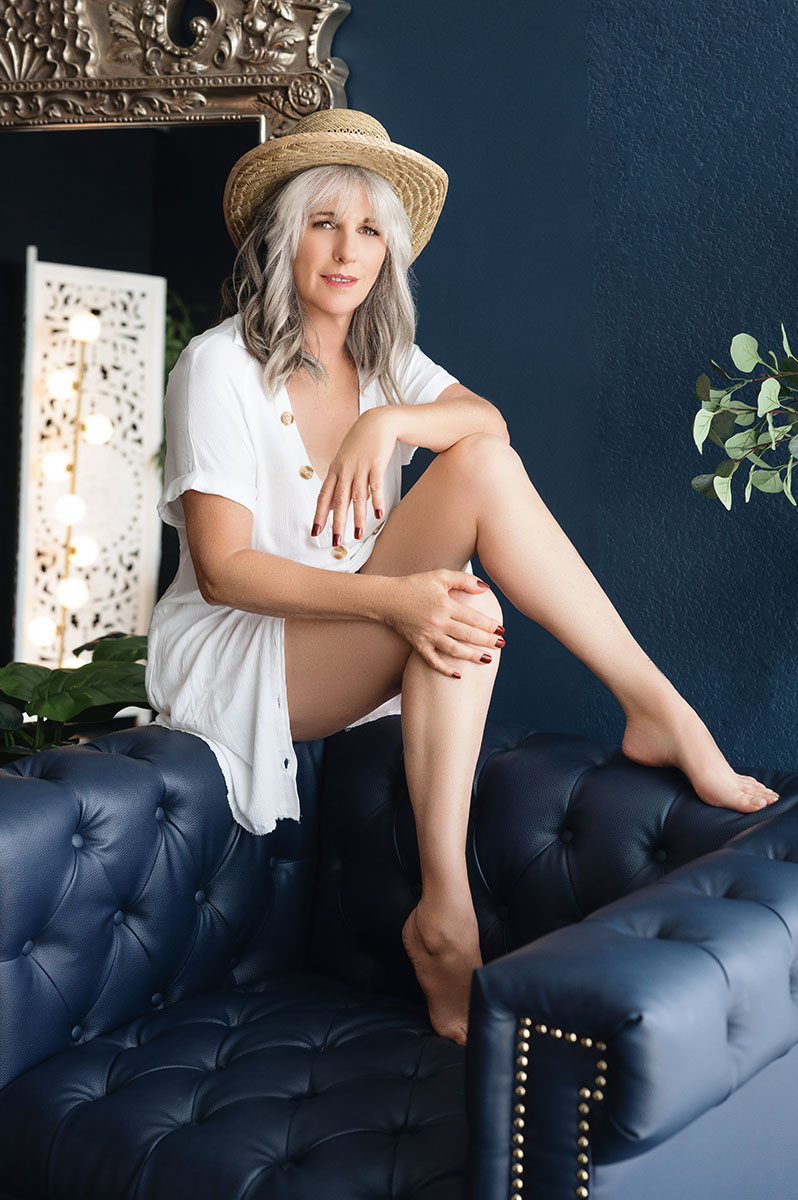 Portrait of mature woman posing sitting on black tufted chair wearing a white dress and a straw hat