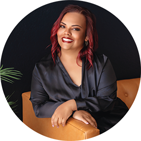 Dark skinned woman with red hair sitting on a burnt orange couch wearing a black silk robe for a boudoir portrait