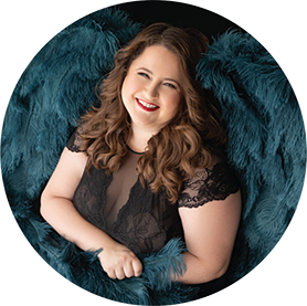 Portrait of curvy woman smiling wearing a sheer black and lace bodysuit and teal wings for a boudoir session