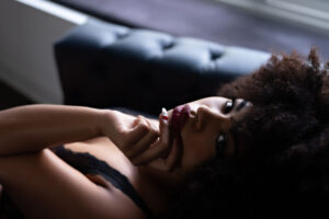 Dark sultry boudoir portrait of bark skinned woman posing on black tufted couch