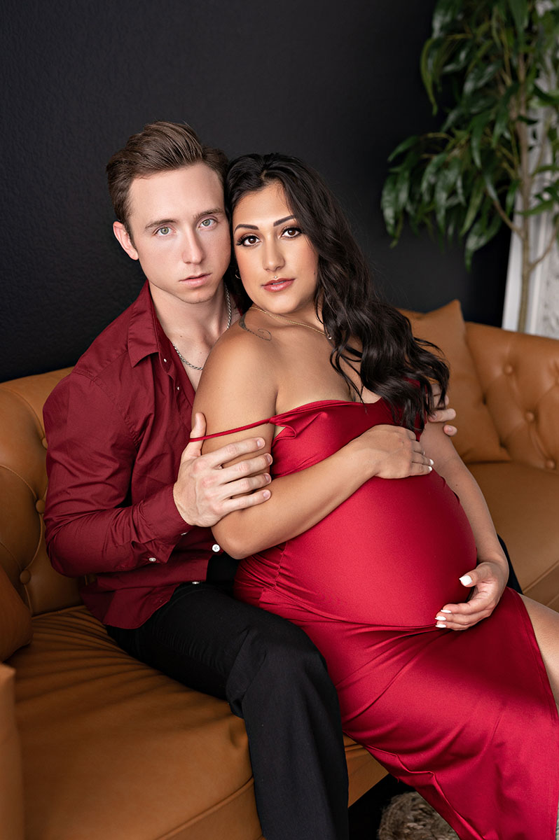 Portrait of a couple posing for a maternity photoshoot where a woman is wearing a long red gown and a an is embracing her wearing a dark red long sleeve. The couple is sitting on a burnt orange couch with a dark wall behind them