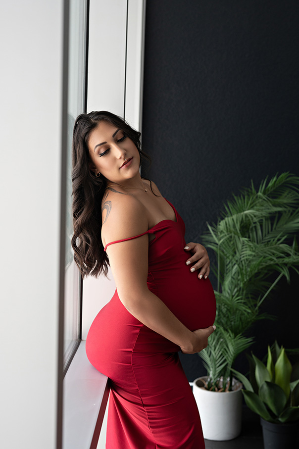 Dimly lit portrait of pregnant woman wearing a red gown leaning against window