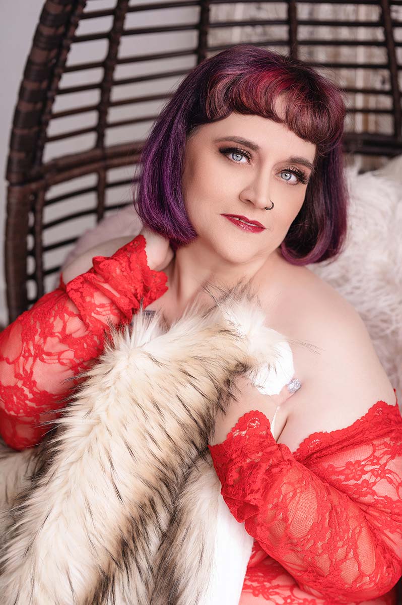 Boudoir portrait of curvy woman with a red and purple bob posing sitting wearing a red lace bodysuit clutching a fur blanket