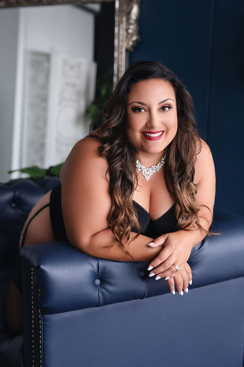 Bright boudoir portrait of curvy brunette woman posing leaning over the edge of a blue tufted couch smiling wearing black lingerie