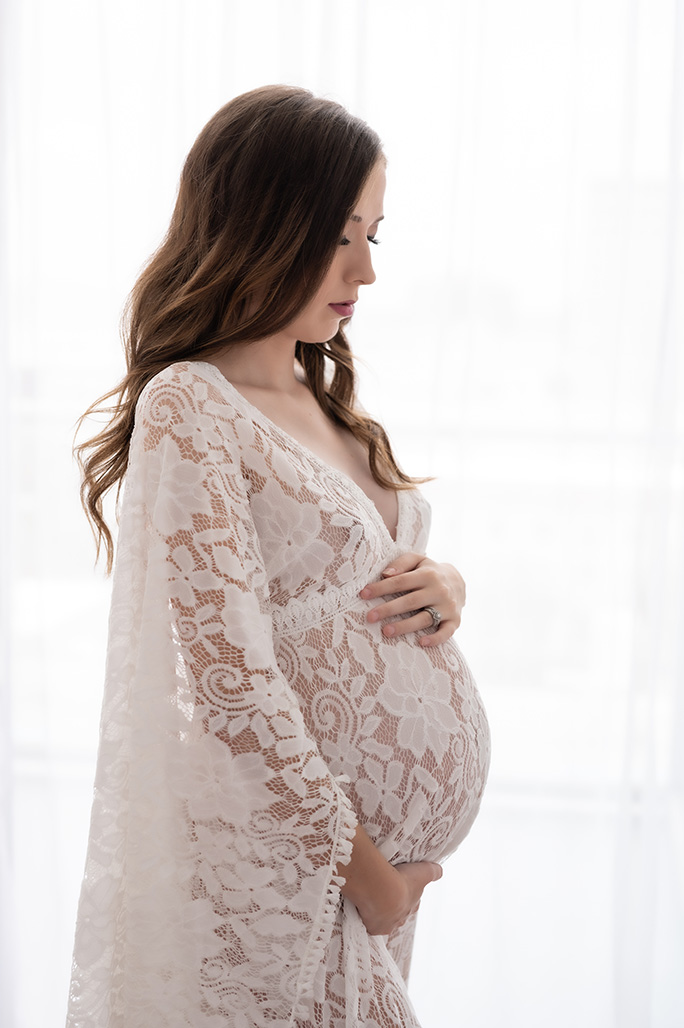 Brightly lit maternity portrait of brunette woman in lace gown holding her belly